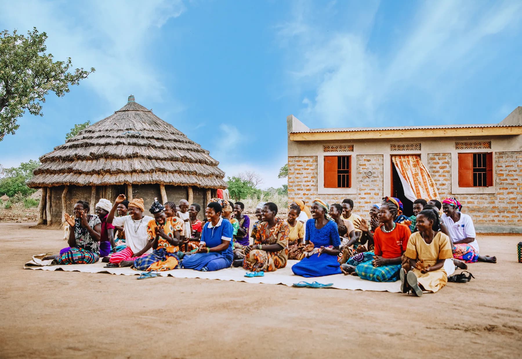 Women from a Homes 4 Homes community sitting together in front of a new brick home and the thatched hut it replaces in Northern Uganda