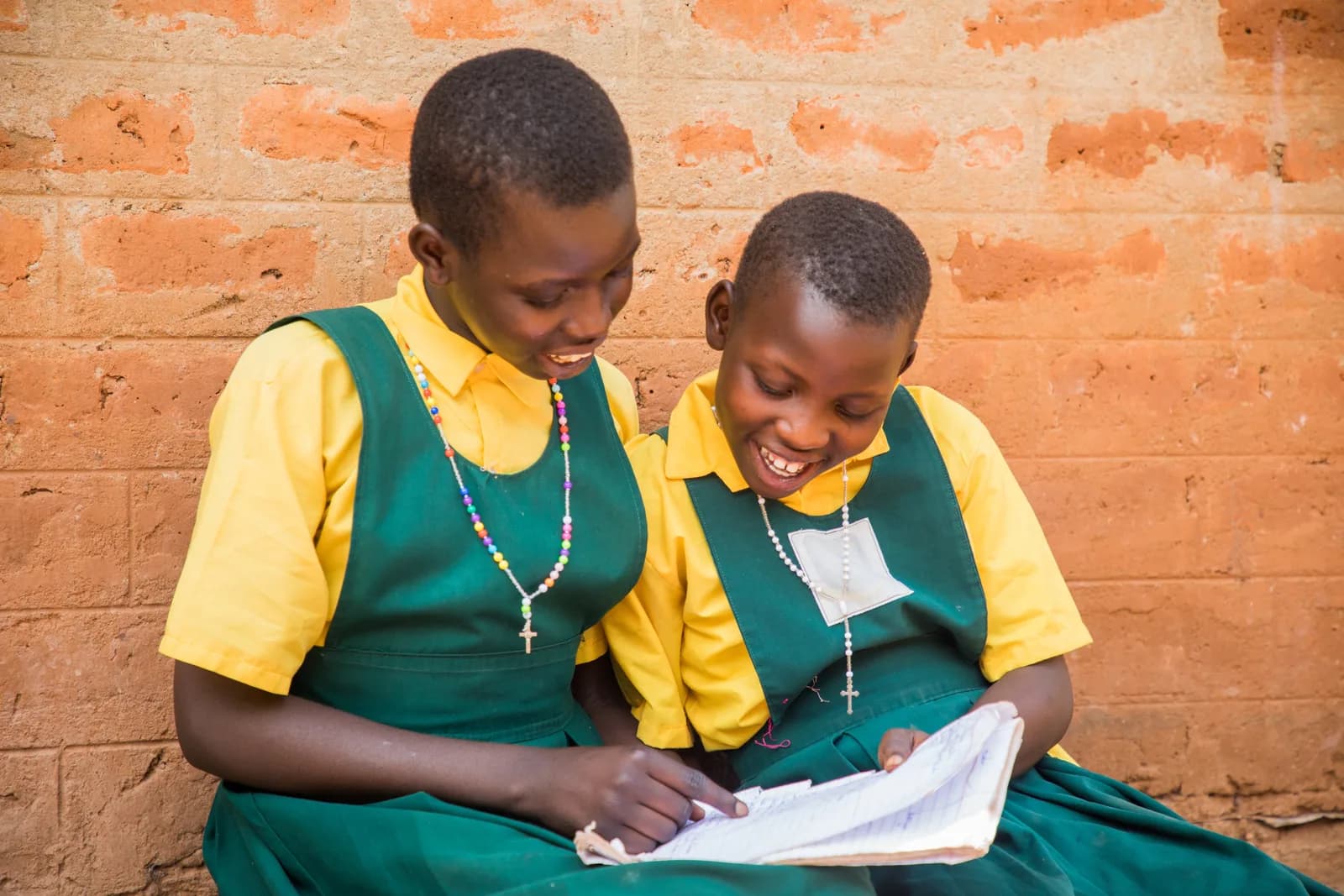 Two schoolgirls in uniform laughing over a notebook against the brick wall of their home