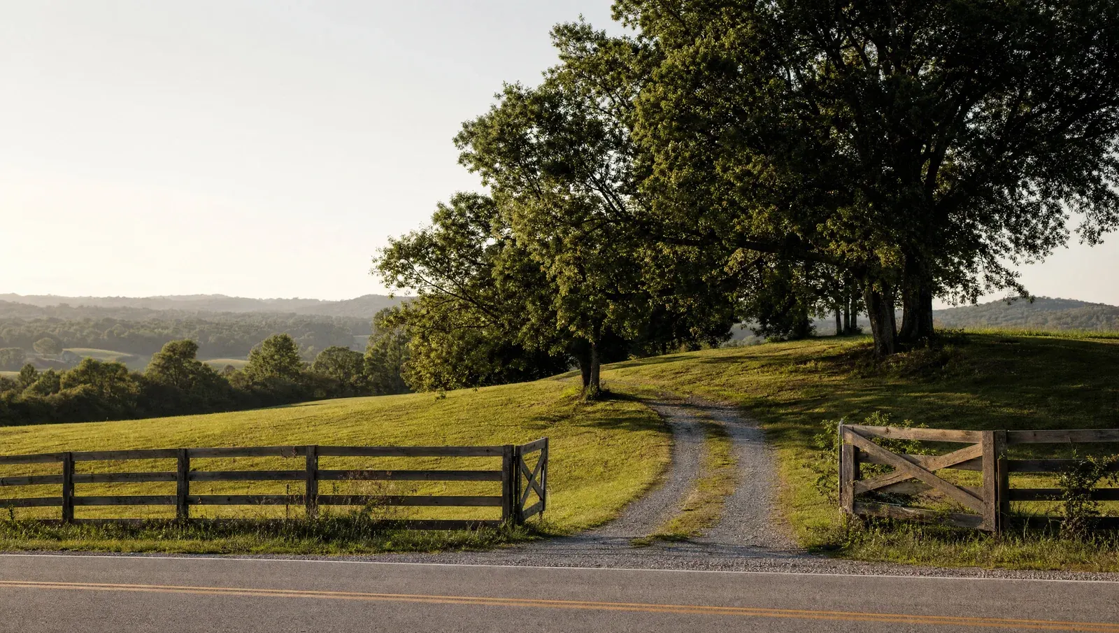 A gravel lane leading from a two-lane country road through an open wooden gate into rolling pasture shaded by old hardwood trees, with hills in the distance