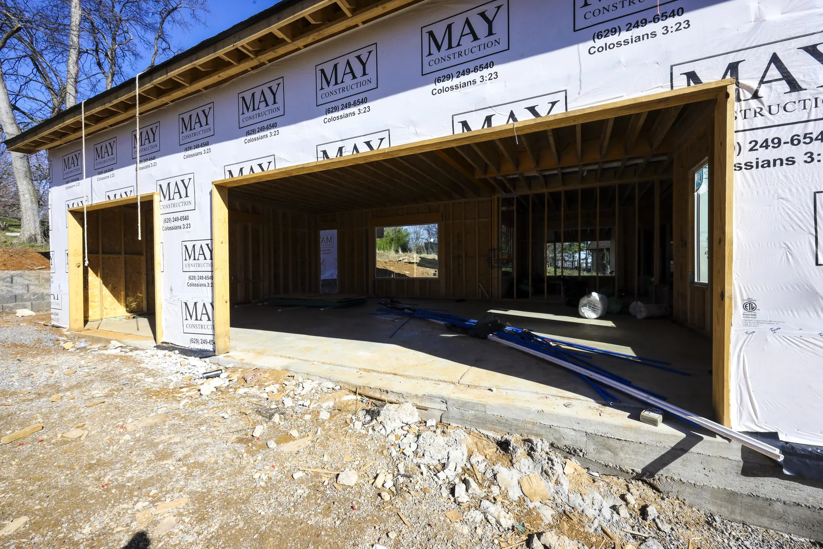 Framed garage bays wrapped in May Construction housewrap, with plumbing pipe and insulation staged on the concrete slab