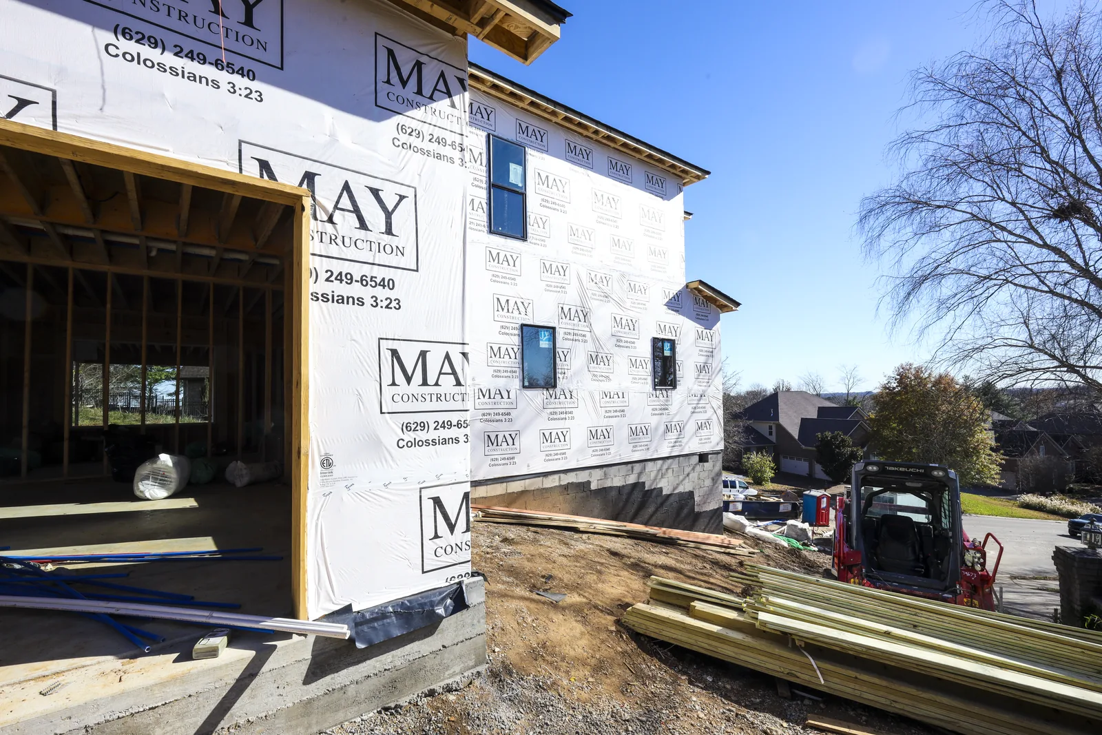 Side elevation of 123 Glen Leven Way in May Construction housewrap, the block foundation stepping down the slope and framing lumber staged in the drive