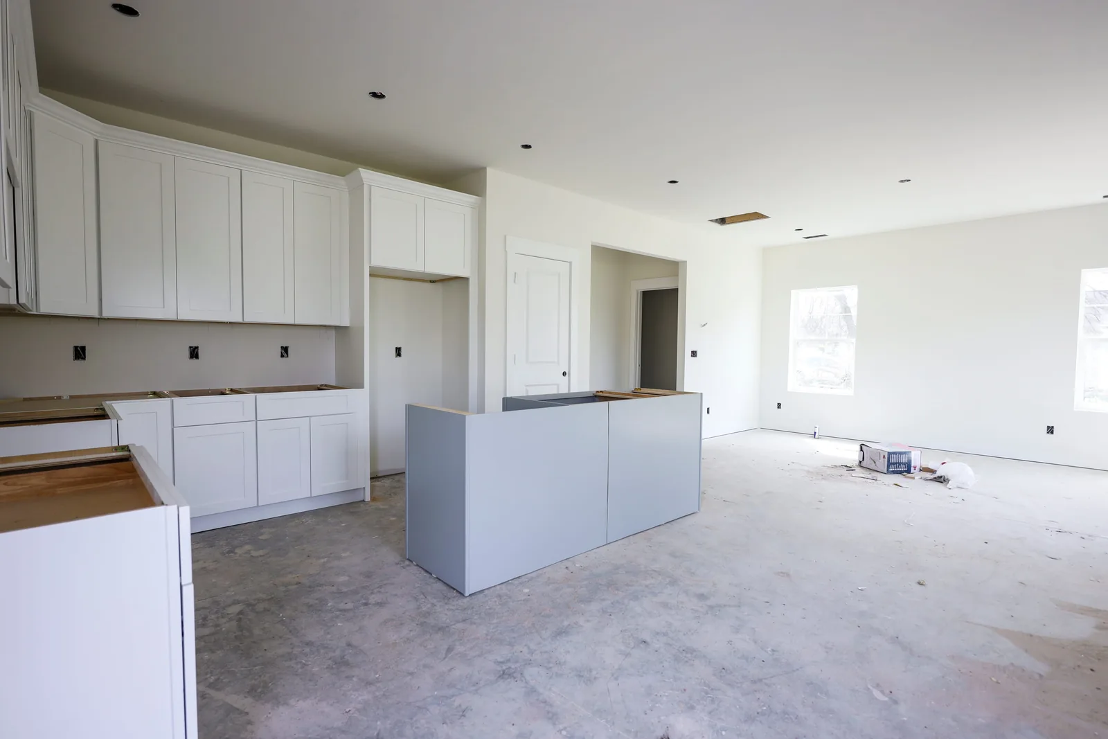 Kitchen mid-install with white shaker cabinets hung, a gray island base in place, and counters not yet set on a bare concrete floor